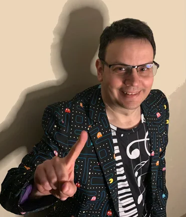 Chris in a Pac-Man print jacket and a piano-key shirt, smiling and holding up one finger